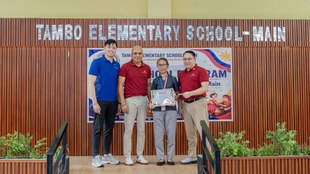 Representatives holding a certificate in front of the Tambo Elementary School main building sign