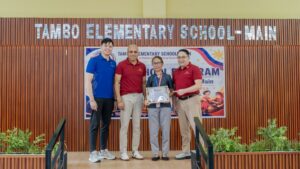 Representatives holding a certificate in front of the Tambo Elementary School main building sign