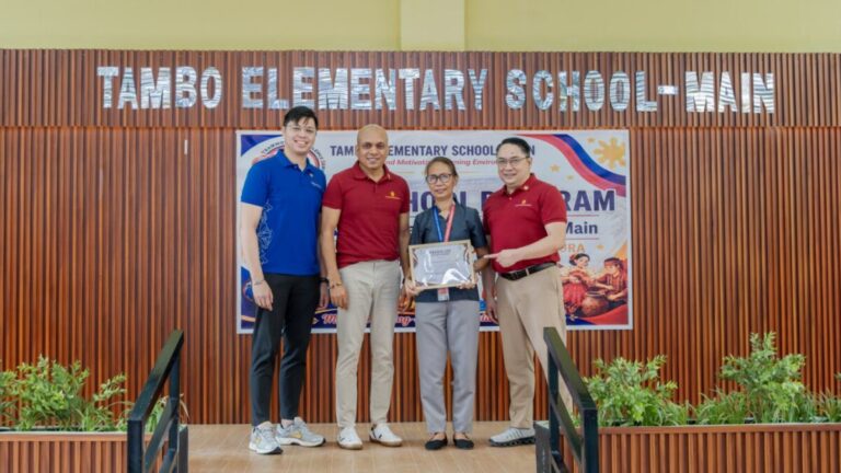 Representatives holding a certificate in front of the Tambo Elementary School main building sign